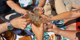 Freunde stoßen beim Picknick an der Isar mit Tumblergläsern an, darin WUYD Single Malt Whisky.