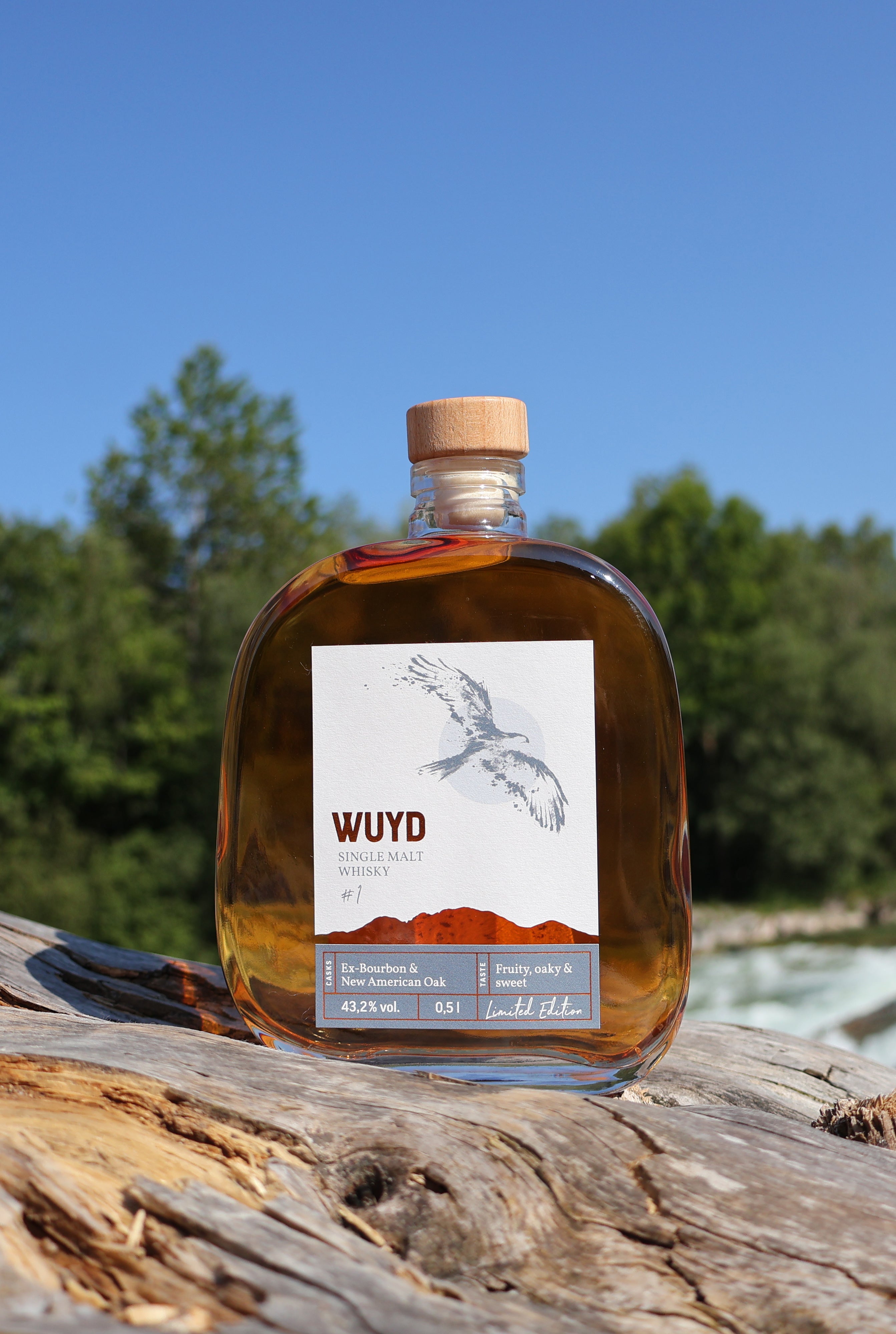 Eine Flasche WUYD Single Malt Whisky steht auf einem Holzstamm, im Hintergrund fließt die Isar.