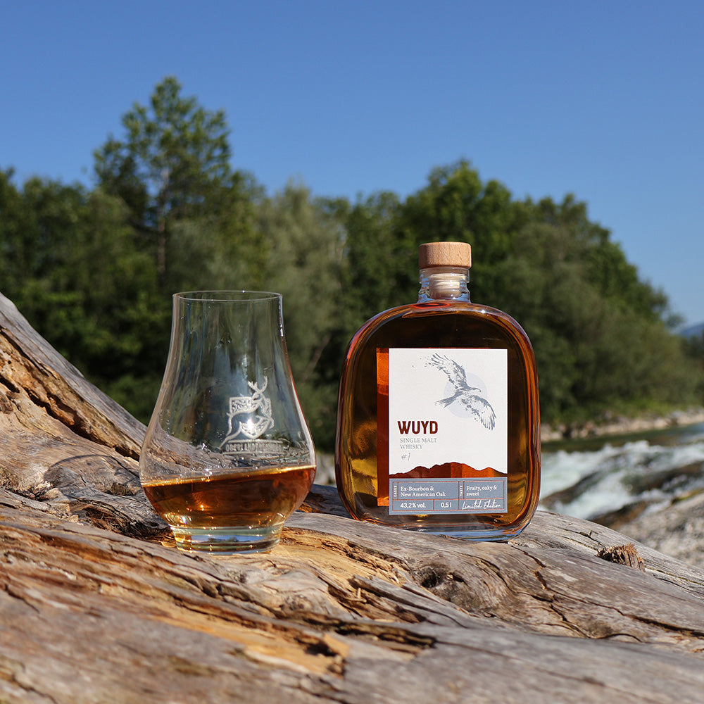 Eine Flasche WUYD Single Malt Whisky und ein Whiskyglas stehen auf einem Holzstamm, im Hintergrund fließt die Isar.