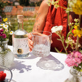 Frau hält ein Tumbler mit Flower Power Gin am Sommertisch. Symbolbild für Drink Garden Glow.