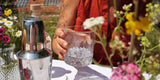 Eine Frau hält ein Glas mit Flower Power Gin, daneben ein Cocktailshaker auf einem sommerlichen Tisch.
