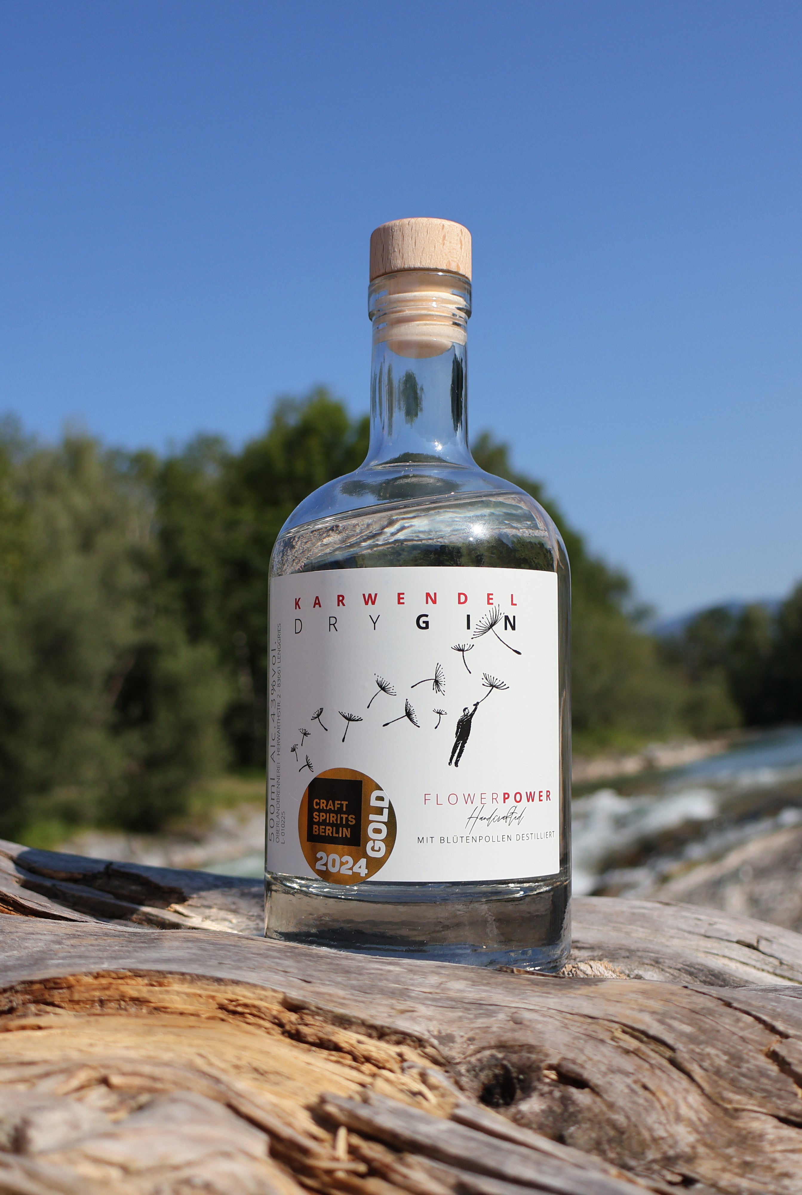 Eine Flasche Flower Power Gin steht auf einem Holzstamm, im Hintergrund fließt die Isar.