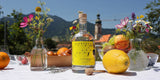 Eine Flasche Flower Power Lemon Gin auf einem sommerlich gedeckten Tisch mit Bergpanorama.