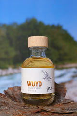 Eine Miniaturflasche WUYD Single Malt Whisky steht auf einem Stück Holz, im Hintergrund fließt die Isar.