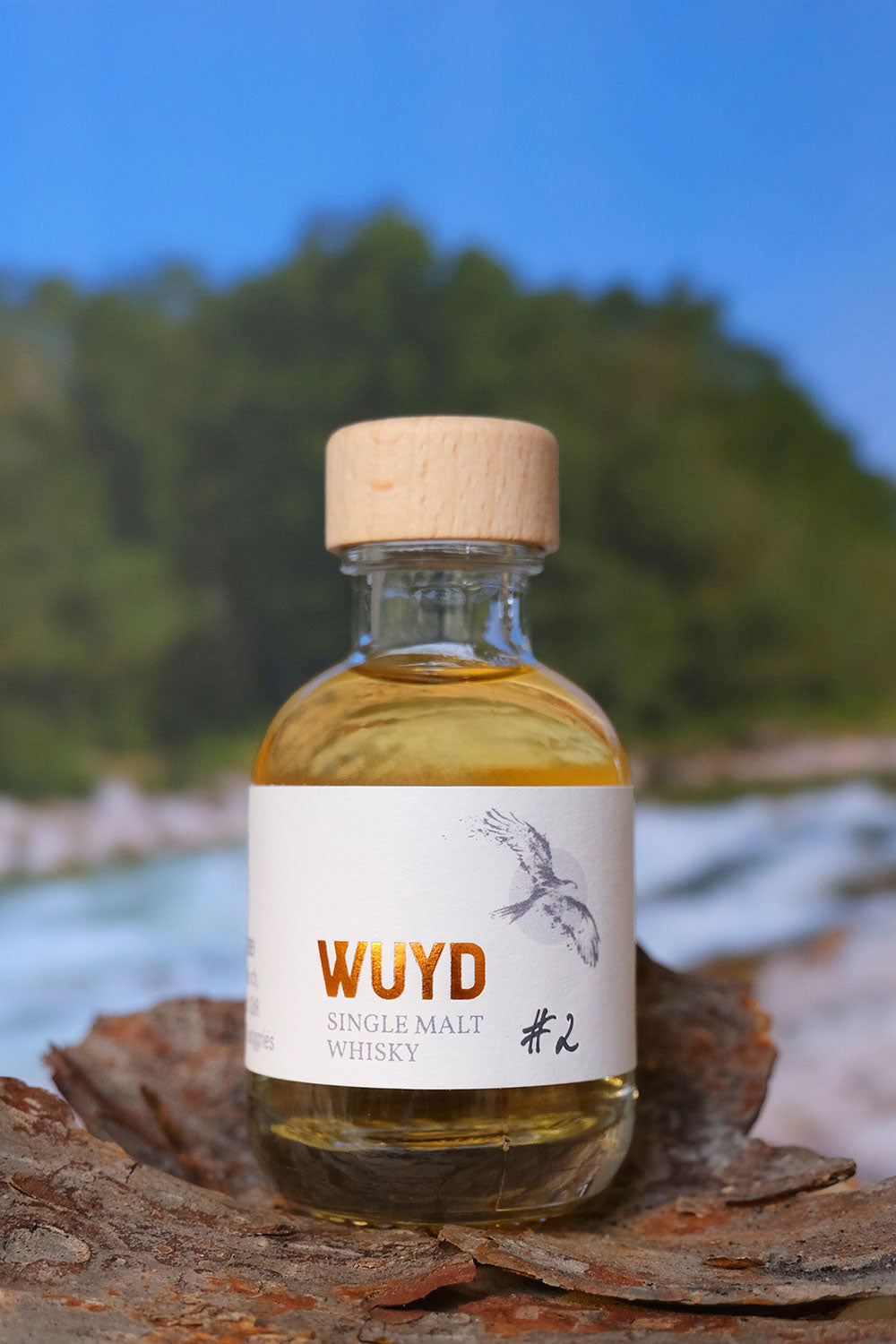 Eine Miniaturflasche WUYD Single Malt Whisky Batch #2 steht auf einem Stück Holz, im Hintergrund fließt die Isar.