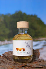 Eine Miniaturflasche WUYD Single Malt Whisky Batch #2 steht auf einem Stück Holz, im Hintergrund fließt die Isar.