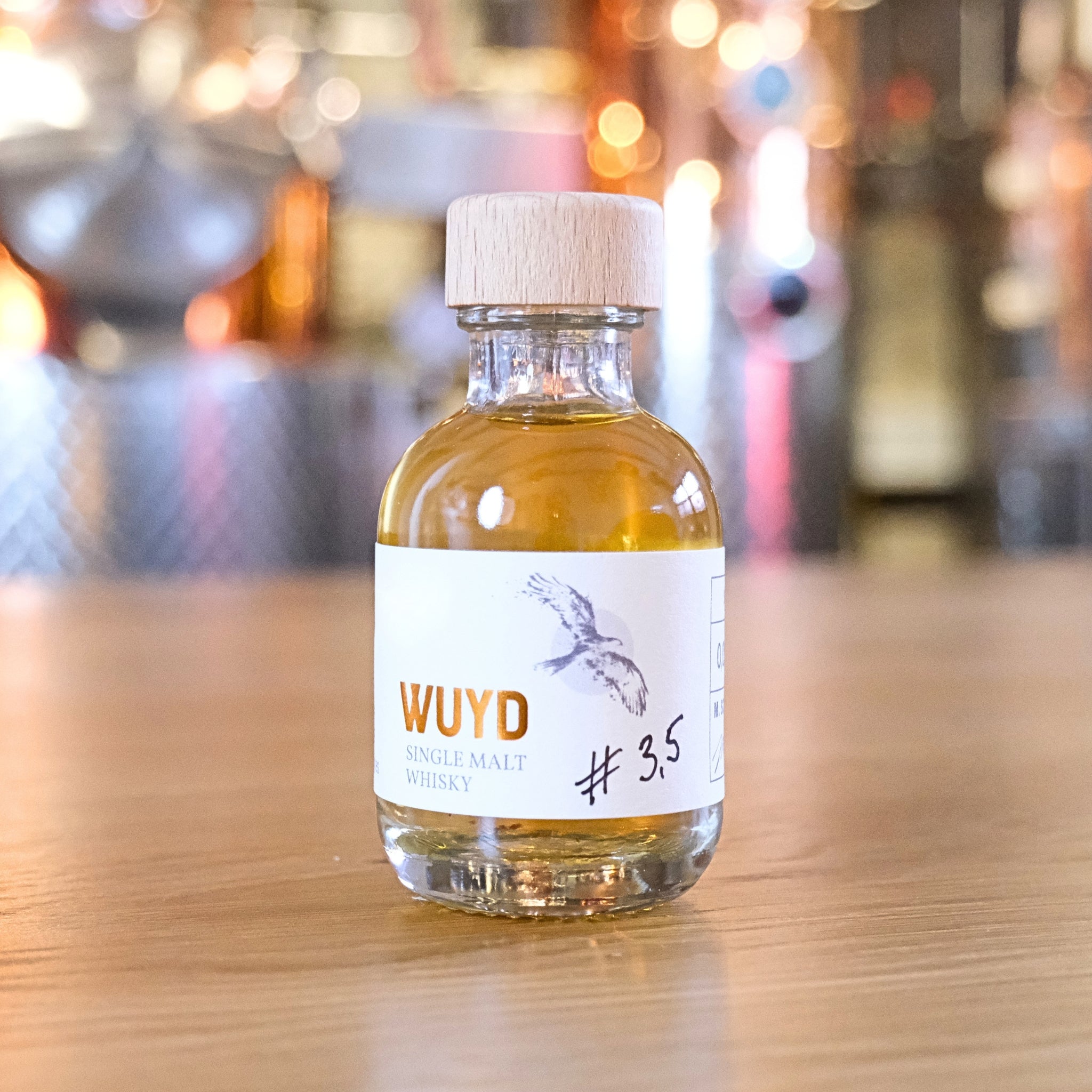 Eine Flasche WUYD Single Malt Whisky Batch #3,5 aus dem Hagebuttenweinfass steht auf einem Tisch, dahinter die kupferne Brennanlage.