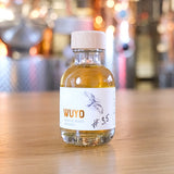 Eine Flasche WUYD Single Malt Whisky Batch #3,5 aus dem Hagebuttenweinfass steht auf einem Tisch, dahinter die kupferne Brennanlage.