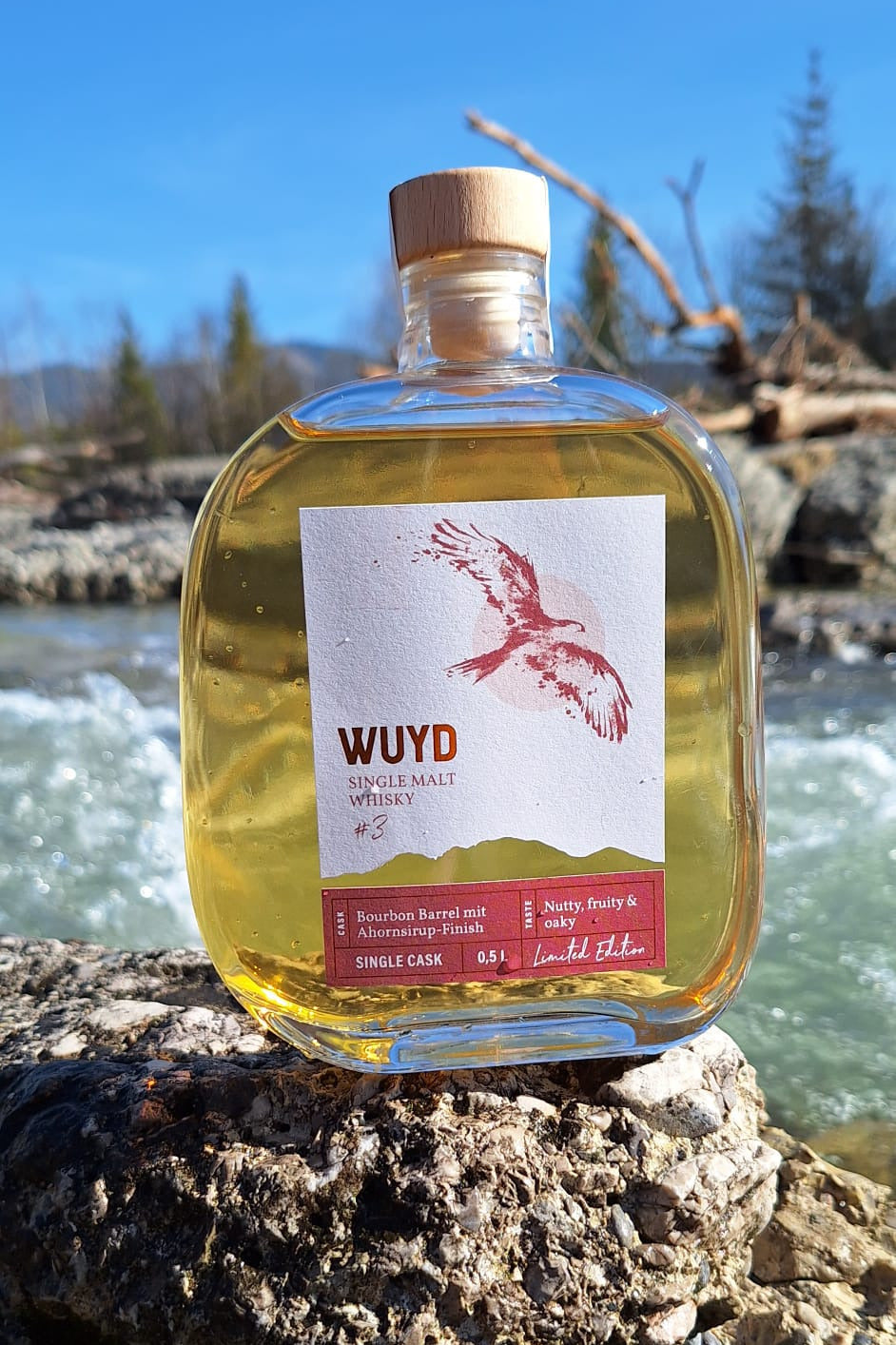 Eine Flasche WUYD Single Malt Whisky Batch #3 steht am Ufer auf einem Stein, im Hintergrund fließt die Isar.
