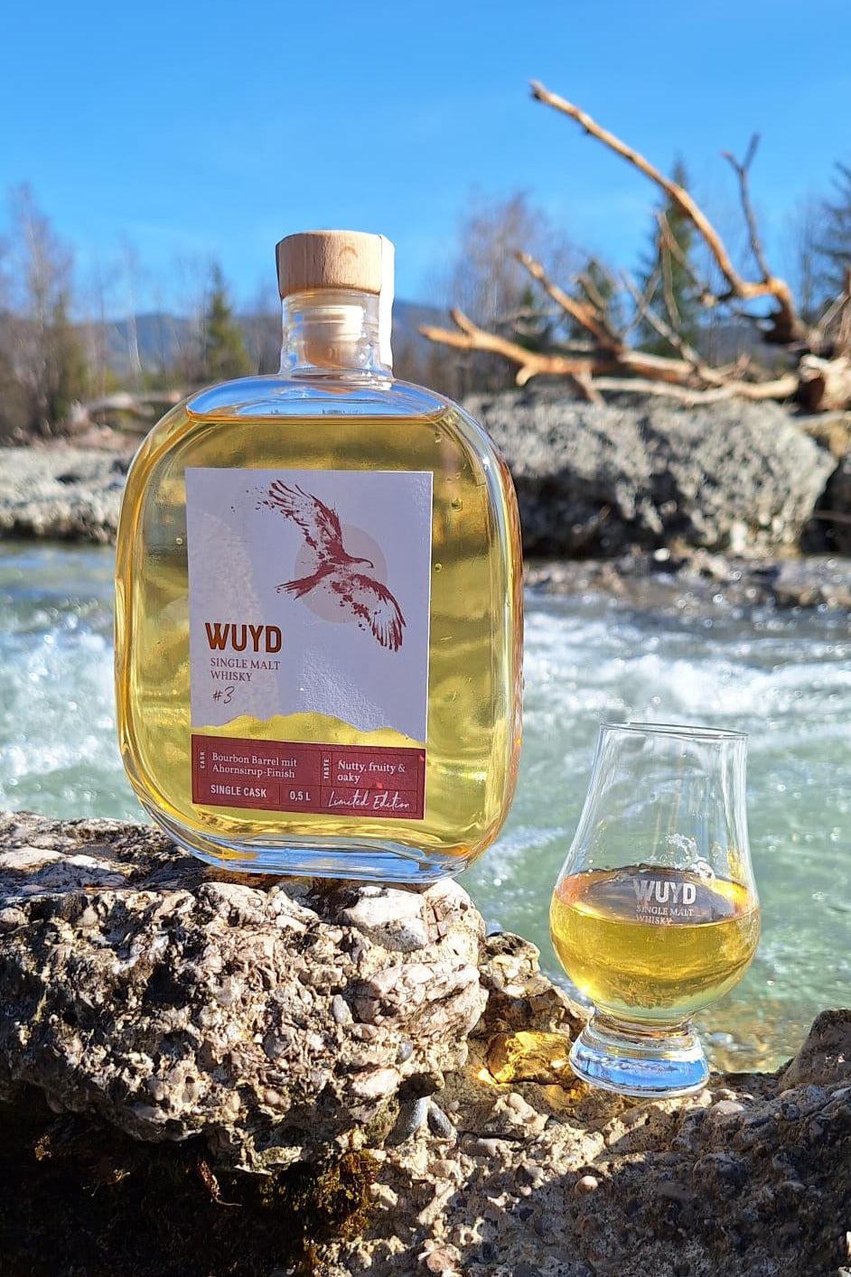 Eine Flasche WUYD Single Malt Whisky Batch #3 und ein Nosingglas mit Whisky stehen am Ufer auf einem Stein, im Hintergrund fließt die Isar.