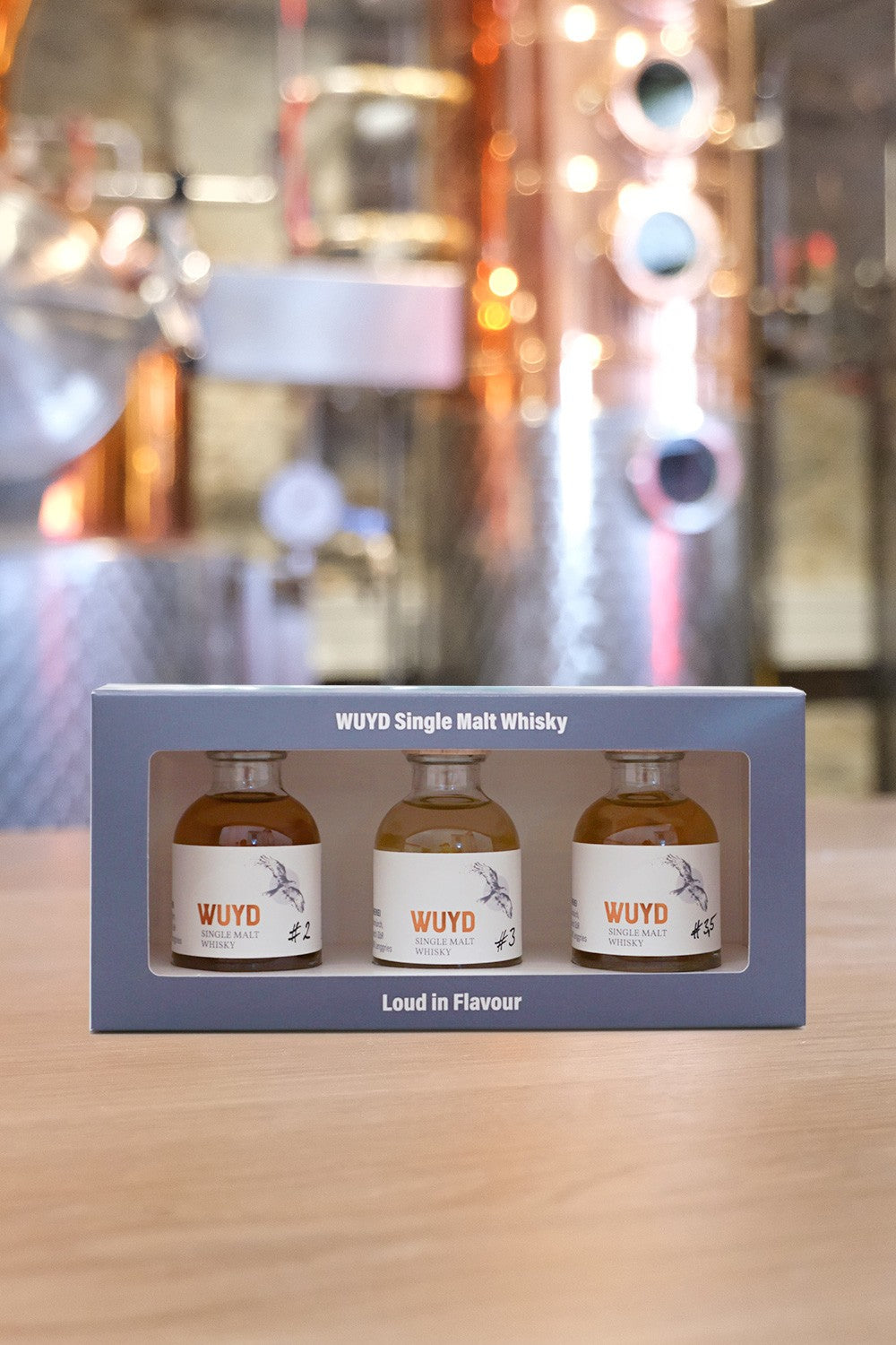 Die Tasting-Box der Oberlandbrennerei  (Vorderseite) mit drei kleinen Flaschen WUYD Single Malt Whisky (Tasting-Set) steht auf einem Tisch, im Hintergrund unscharf die Brennblase.