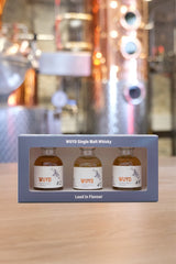 Die Tasting-Box der Oberlandbrennerei  (Vorderseite) mit drei kleinen Flaschen WUYD Single Malt Whisky (Tasting-Set) steht auf einem Tisch, im Hintergrund unscharf die Brennblase.