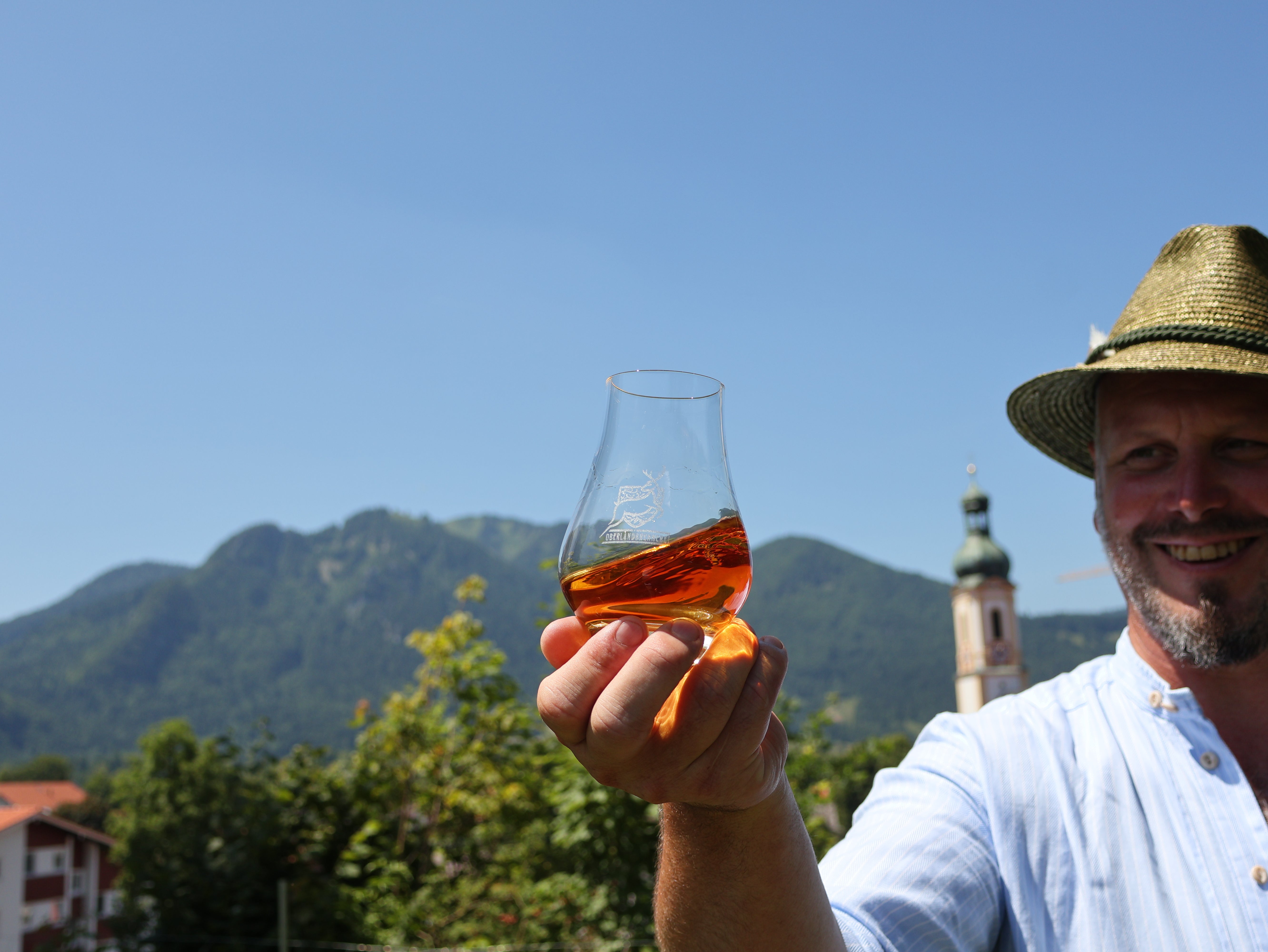 Ein Mann mit einem Hut hält vor Bergkulisse ein Nosingglas mit Whisky in seiner Hand.
