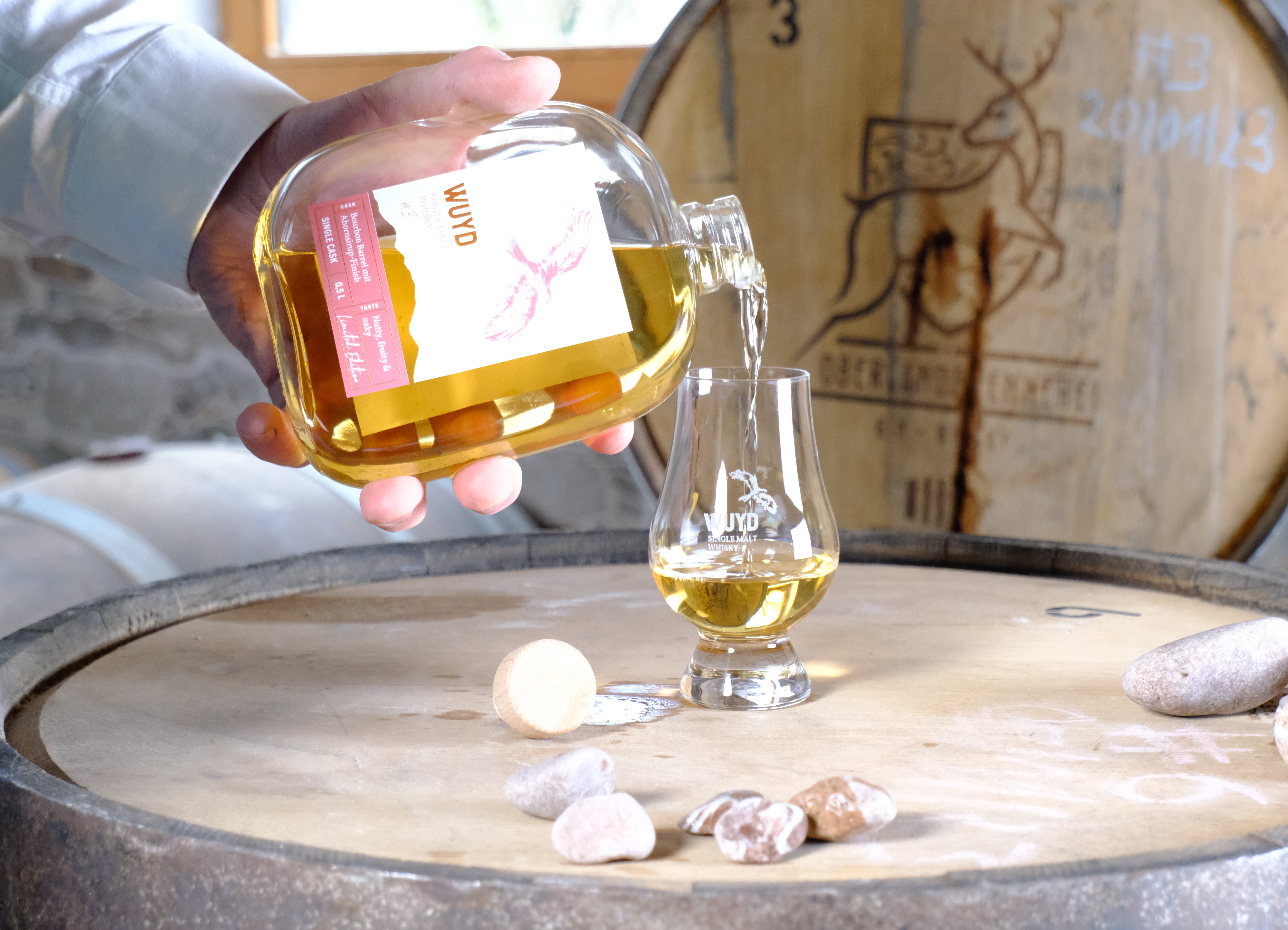 Eine Hand hält eine Flasche WUYD Single Malt Whisky und gießt daraus Whisky in ein Nosingglas, das auf einem Whiskyfass steht.