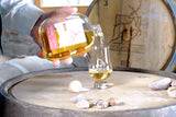 Eine Hand hält eine Flasche WUYD Single Malt Whisky und gießt daraus Whisky in ein Nosingglas, das auf einem Whiskyfass steht.