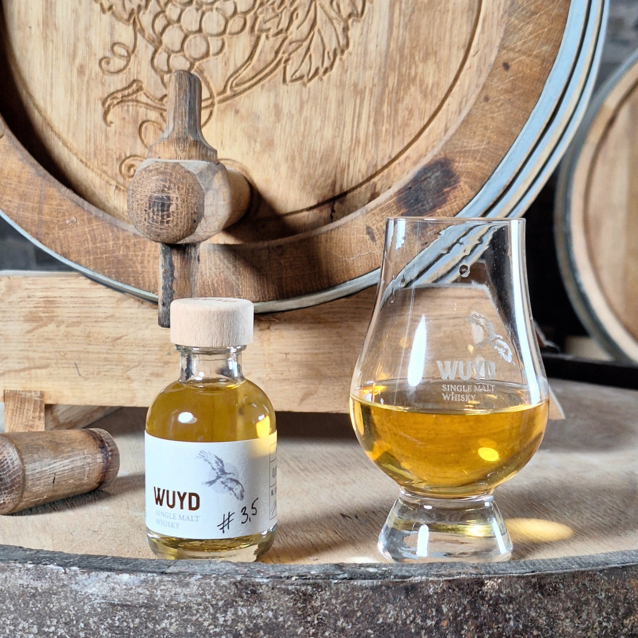 Eine Flasche WUYD Single Malt Whisky Batch #3,5 aus dem Hagebuttenweinfass steht neben einem Whiskyglas auf einem Fass, dahinter das Hagebuttenweinfass.