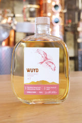 Eine Flasche WUYD Single Malt Whisky Batch #3 steht auf einem Holztisch, im Hintergrund ist verschwommen eine Brennblase zu sehen.