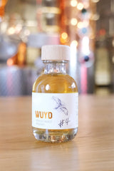Eine Flasche WUYD Single Malt Whisky Batch #4 aus dem Sherryfass steht auf einem Tisch, dahinter die kupferne Brennanlage.