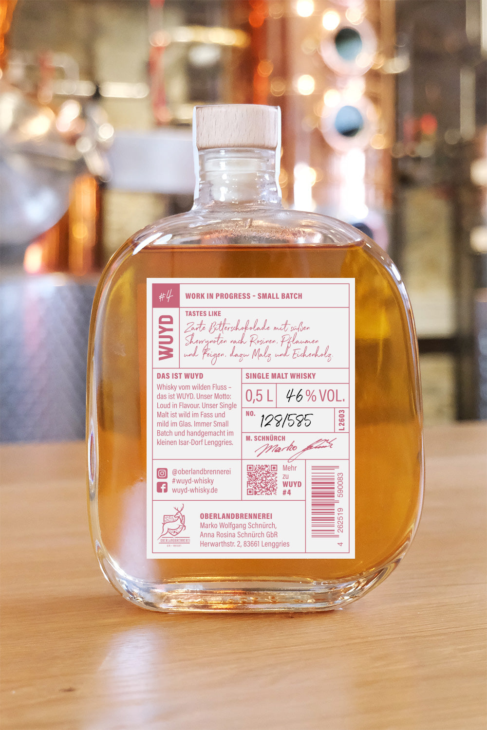 Eine Flasche WUYD Single Malt Whisky Batch #4 (Rückseite mit Rücketikett) steht auf einem Holztisch, im Hintergrund ist verschwommen eine Brennblase zu sehen.