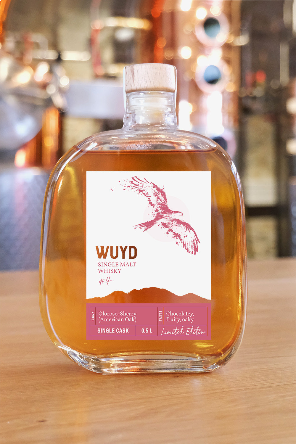 Eine Flasche WUYD Single Malt Whisky Batch #4 (Vorderseite) steht auf einem Holztisch, im Hintergrund ist verschwommen eine Brennblase zu sehen.
