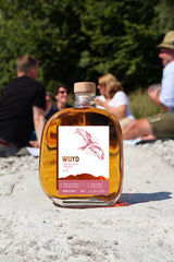 Eine Flasche WUYD Single Malt Whisky Batch 4 steht auf einem Stein am Ufer der Isar, im Hintergrund sitzen Personen.