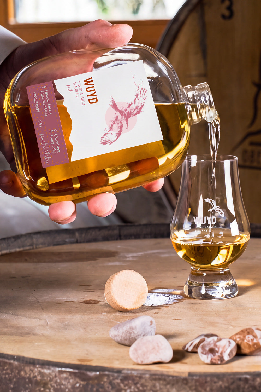 Eine Hand hält eine Flasche WUYD Single Malt Whisky und gießt daraus Whisky in ein Nosingglas, das auf einem Whiskyfass steht.