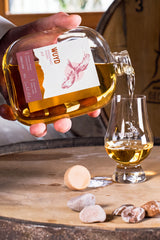 Eine Hand hält eine Flasche WUYD Single Malt Whisky und gießt daraus Whisky in ein Nosingglas, das auf einem Whiskyfass steht.