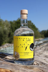 Eine Flasche Flower Power Lemon Gin steht auf einem Holzstamm, im Hintergrund fließt die Isar.